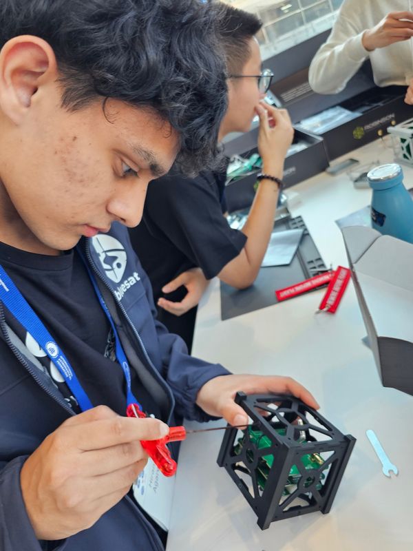Hiranya in a BlueSat jacket fastening a screw into a black 1U CubeSat structure with a red screwdriver, circuit boards visible inside.