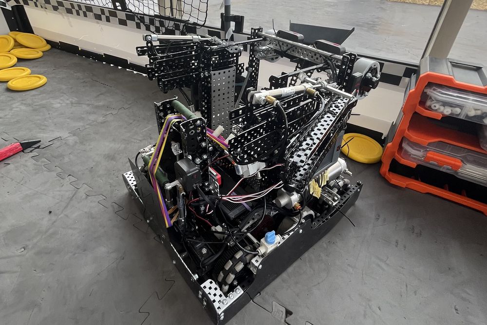 An earlier robot beside a home practice field, with pneumatic cylinders and a folding arm visible and scoring discs stacked on the floor nearby.