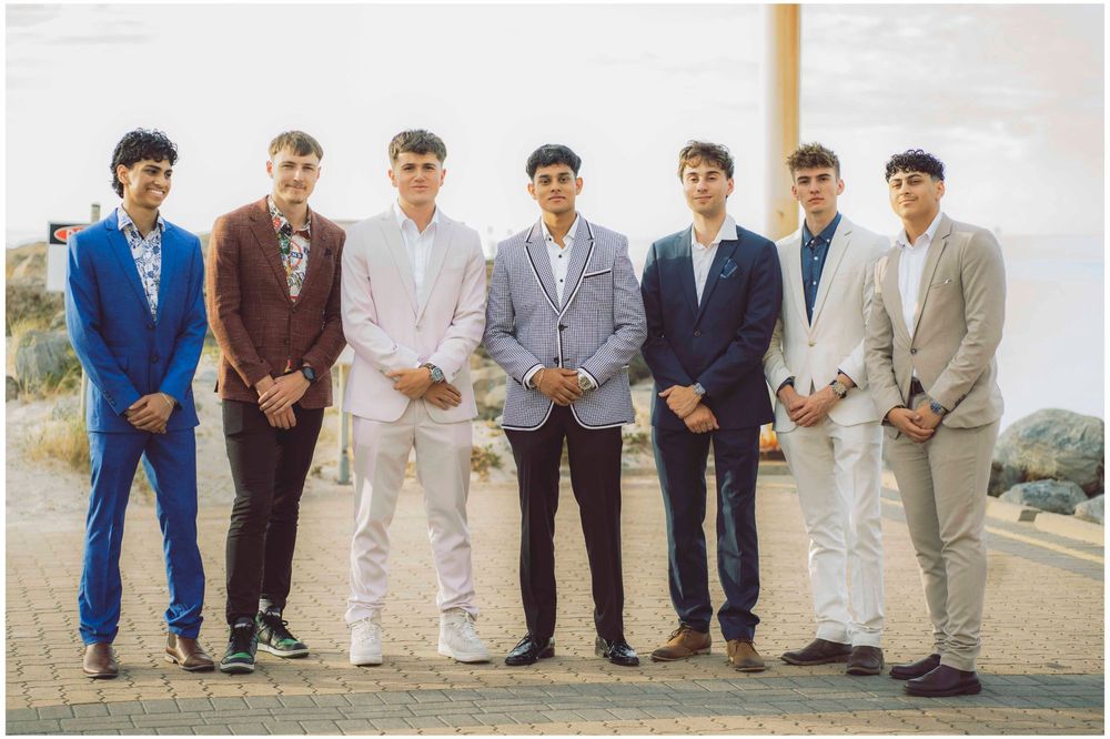 The seven team members in suits standing in a row on a beachfront path, Hiranya in the centre in a checked jacket.